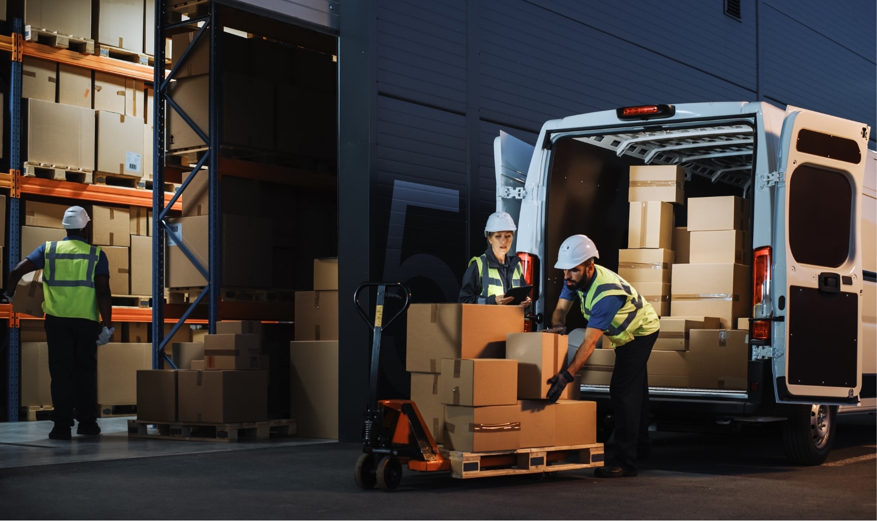 Two logistics operators load parcels into a commercial vehicle outside a warehouse.