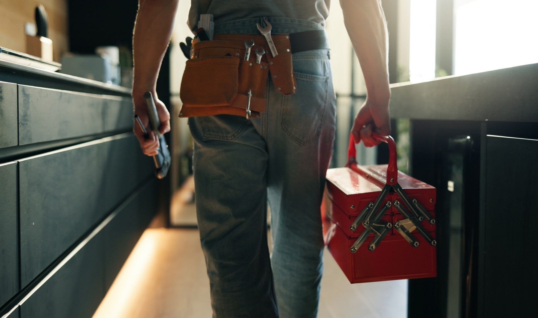 Plombier avec une boîte à outils, vu de dos, intervenant au domicile d’un client pour un entretien, une réparation ou une inspection de fuite dans une cuisine. Équipement professionnel, service client et intervention de dépannage en plomberie.